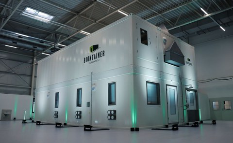 White containers form a modular process plant. This stands in a hall and is partially illuminated in green.