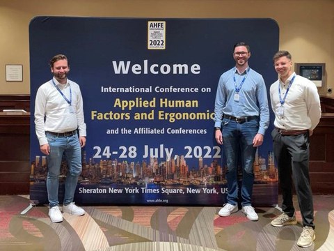 Three men in formal dress stand in front of a sign reading AHFE 2022