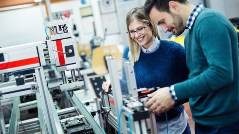 Das Foto zeigt zwei Studierende, die an einer hochtechnisierten Maschine stehen. Die Studierenden lächeln.