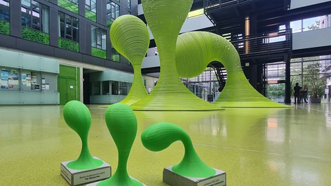Im Hintergrund sind die tropfenförmigen grünen Stahlskulpturen aus dem Foyer, im Vordergrund sind Miniaturausgaben dieser Skulpturen auf Betonsockeln mit Aufschriften der Preistragenden.