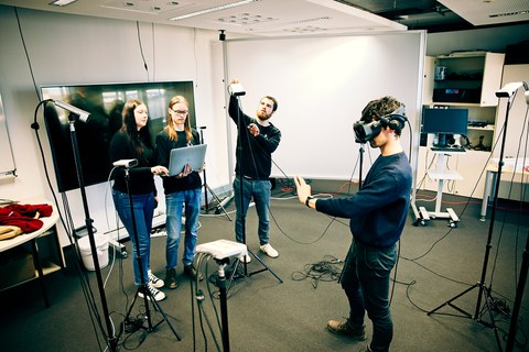 Vier Studierende beim Experimentieren im Computergrafik-Labor, einer mit VR-Brille im Kreis mit Kameras, zwei mit Laptop seine Bewegungen prüfend, einer beim Einstellen einer Kamera