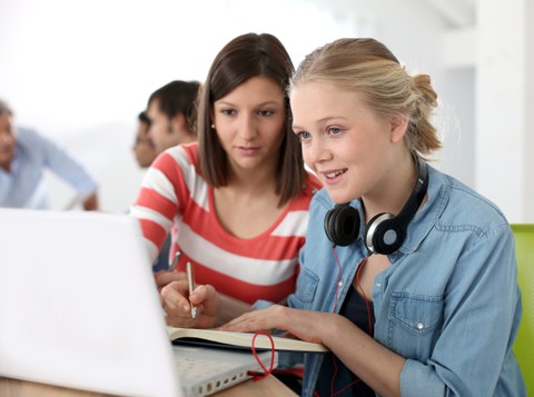 Das Foto zeigt zwei Studentinnen, die gemeinsam auf einen Laptop schauen und sich etwas in ein Heft notieren. 