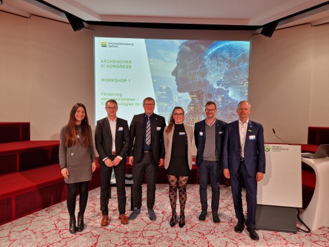 Im Vordergrund stehen 6 Personen, im Hintergrund ist eine Präsentationsfolie zu sehen.