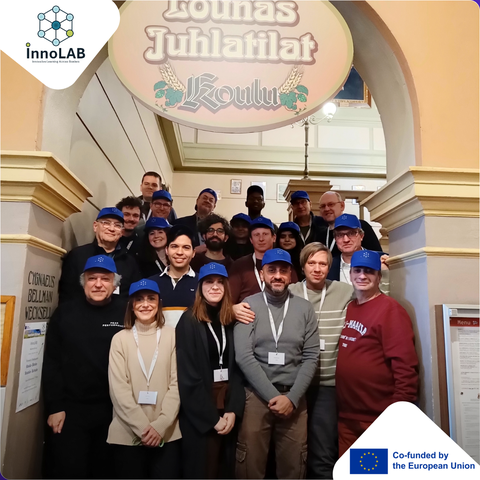 Now translate to German: A group of about twenty people wearing matching blue caps stands together on an indoor staircase beneath a decorative sign. Most are smiling and wearing name badges.
