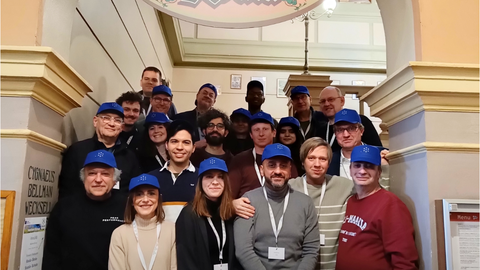 Eine Gruppe von etwa zwanzig Personen mit passenden blauen Caps steht gemeinsam auf einer Innentreppe unter einem dekorativen Schild. Die meisten lächeln und tragen Namensschilder. 