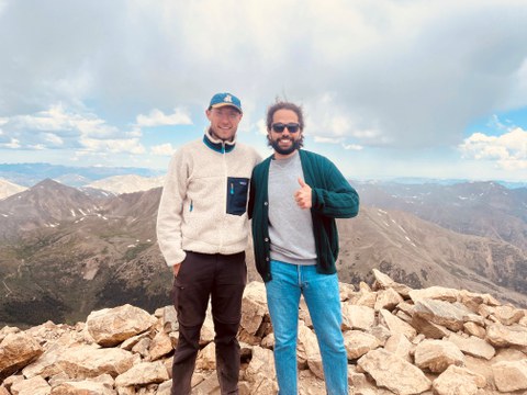 Zwei Männer, Herr Guesmi und Herr Manthey, in Wanderkleidung auf einem Berggipfel, zufrieden lächelnd.