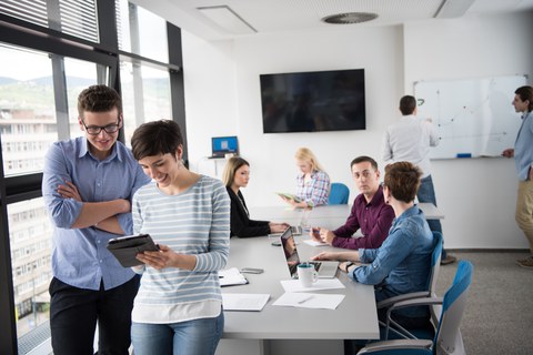 Foto von acht Menschen in einem Büro, zwei von ihnen sind im Vordergrund und schauen auf ein Tablet. Die anderen sitzen sprechend an einem Tisch beziehungsweise stehen vor einem Whiteboard.