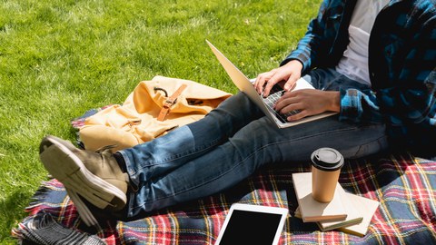 Das Foto zeigt eine studierende Person mit Laptop im Anschnitt. In der Nähe liegen ein Tablet mit leerem Bildschirm und Pappbecher auf einer karierten Decke.