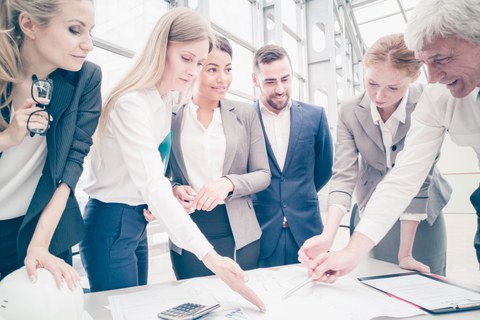 Das Foto zeigt mehrere Personen in Businesskleidung, die gemeinsam um einen Tisch herum stehen und verschiedene Zettel und Pläne betrachten.