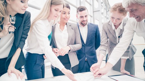 Das Foto zeigt mehrere Personen in Businesskleidung, die gemeinsam um einen Tisch herum stehen und verschiedene Zettel und Pläne betrachten.
