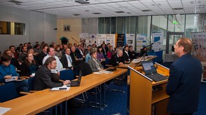 The image depicts the auditorium at Fraunhofer IKTS. Almost all seats are occupied by guests, listening with interest to a scientific lecture. A number of scientific posters are presented in the back of the room.