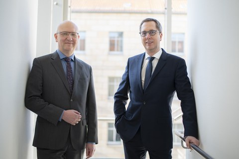 Two men stand in front of a window and smile at the camera.