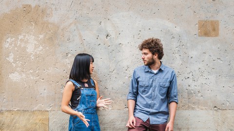 Photo: An Asian woman and a man lean against a concrete wall and talk. 