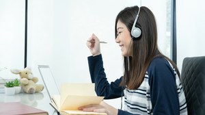 The photo shows a young woman at her desk. She is wearing headphones, smiling and writing something down in her notebook. 