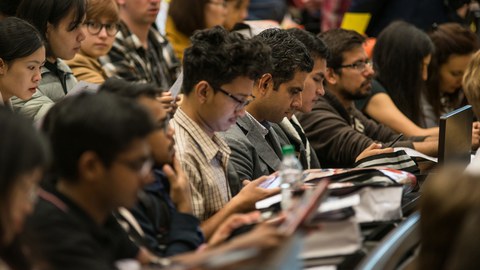 The photo shows the well-filled rows of benches in a lecture hall. Many of the students are looking at their mobile phones or at the bags in front of them.
