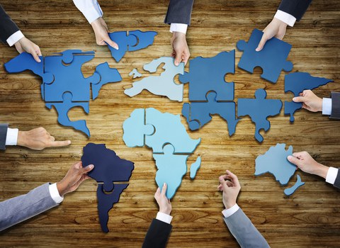 The photo shows the hands of people in business clothes. They are standing around a table. Pieces of a puzzle are lying on the table. Put together they form a map of the world.