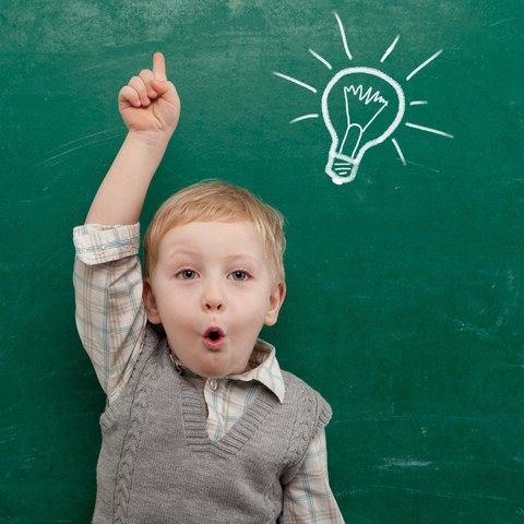Child stretches his finger upwards and answers. In the background is a blackboard with a light bulb painted in chalk.
