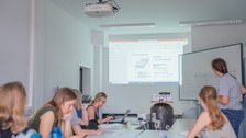 Im Hintergrund: Beamer mit Präsentation, im Vordergrund: junge Frauen an einem Tisch.