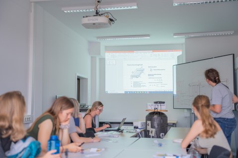 Im Hintergrund: Beamer mit Präsentation, im Vordergrund: junge Frauen an einem Tisch.