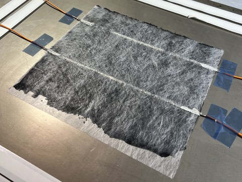 Auf einer Metallplatte befindet sich eine Folie. Darin eingebettet sind jeweils zwei Kabel, die mit Klebeband an der Metallplatte befestigt sind.