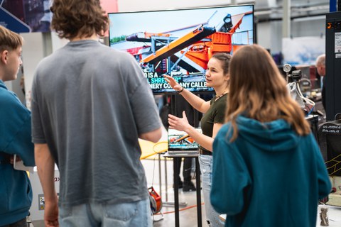 A young woman stands in the middle of a group of young people and gesticulates as she explains. A screen with a machine can be seen in the background. The young people are standing with their backs to the viewer.
