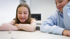 Two girls are sitting at a table, grinning and looking at two spinning tops that are moving on the table.