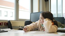 A girl is sitting at a table, one elbow resting on the table. She holds a pen in her other hand and a sheet of paper in front of her. The girl is looking intently out of the window, as if she is thinking hard.