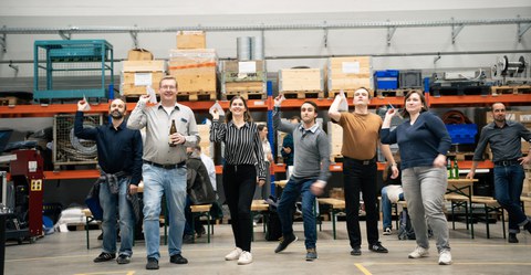 6 people stand next to each other and hold a paper aeroplane in their right hand, which they are about to make fly. They look concentrated. Some are laughing.
