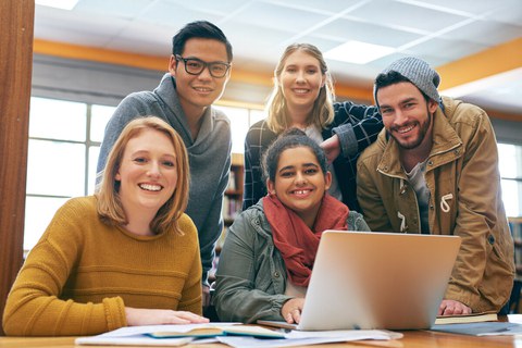 Photo of a group of 5 students studying together in the library and smiling happily at the camera.