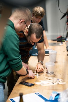 Two people stand concentrated at a table and bend over wooden sticks that they have just fixed in such a way that they form a diamond pattern.