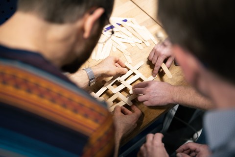 Close-up: two people to the left and right of the image section, joining wooden sticks to form a diamond pattern. Close-up of hands and wooden sticks.