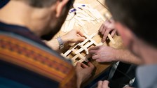 Close-up: two people to the left and right of the image section, joining wooden sticks to form a diamond pattern. Close-up of hands and wooden sticks.