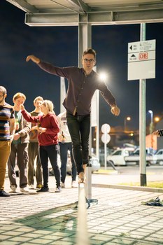 A student is concentrating on a slackline and balancing. It's outside, already nighttime, and other students and staff are standing in the background.