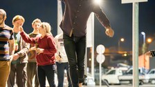 A student is concentrating on a slackline and balancing. It's outside, already nighttime, and other students and staff are standing in the background.