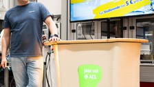 A person stands leaning against a platform. A screen integrated into the platform displays the words ‘Join us @ ACL’.