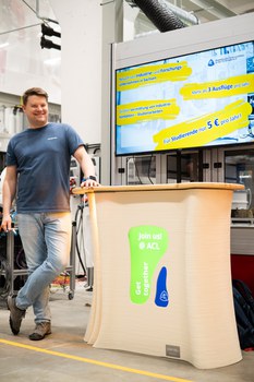 A person stands leaning against a platform. A screen integrated into the platform displays the words ‘Join us @ ACL’.