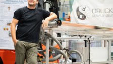 A man leans against an appliance. The rectangular device has many tubes. Glass tubes are attached to the upper side.