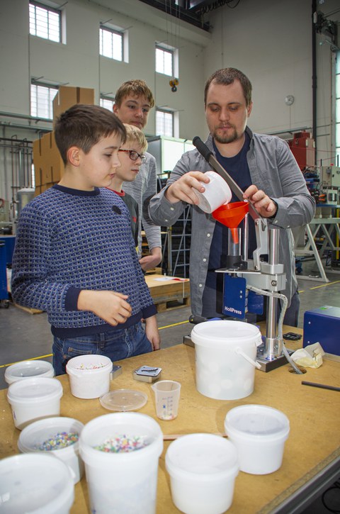 Three boys aged between 11 and 13 stand in front of the HoliPress. An engineer fills granules into a funnel.