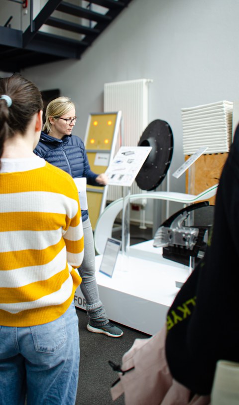 A woman stands in the center of the picture and explains something. Various devices and demonstrators can be seen in the background. In the foreground (left), a girl stands blurred with her back to the viewer.