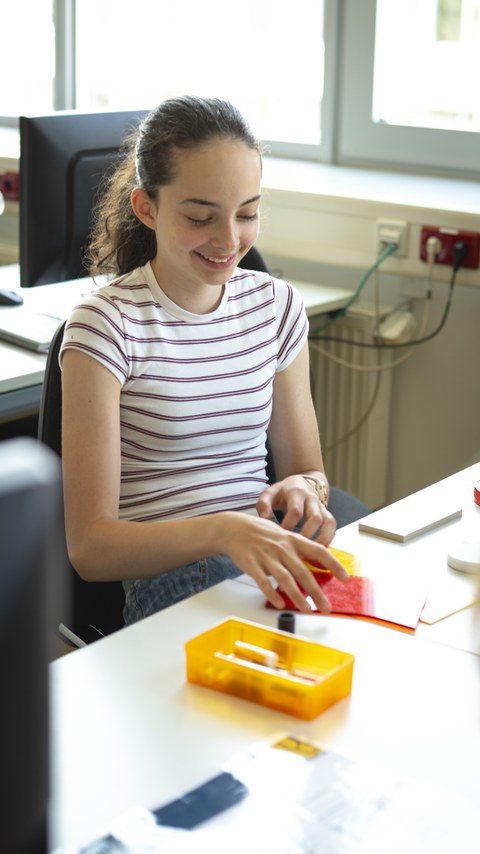 A student is seated at a table, smiling while holding a plastic component in her hand.