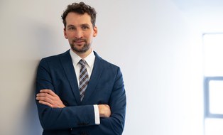 A portrait of a man with black curls and a blue suit. He is leaning against a wall and smiling warmly at the camera.