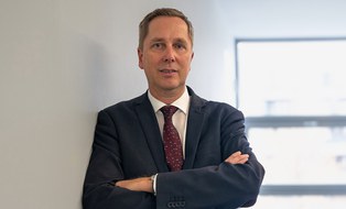 A man in a suit is leaning against a wall and looking at the camera.
