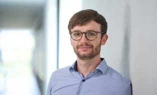 Portaitfoto. Ein Mann mit dunkelblonden Haaren, Pony und Brille lehnt an einer Wand und schaut lächelnd in die Kamera.