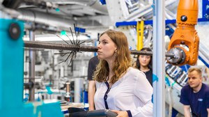 Eine Studentin steht am Flechtrad und hält einen Display in der Hand. Sie schaut prüfend in die Richtung des Flechtrades. Andere Student:innen und ein Techniker stehen im Hintergrund und beobachten sie.