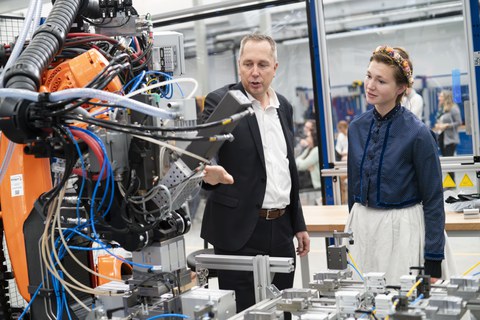 Prof Maik Gude can be seen in the foreground talking to a Sorbian woman in a traditional costume. The two are standing between machines.