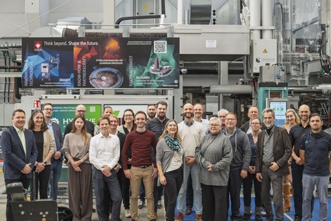 The group photo shows a group of people standing in front of a large machine and looking into the camera. A positive, smiling gesture can be observed.