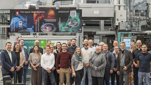Das Gruppenfoto zeigt eine Gruppe von Personen, die vor einer groß dimensionierten Maschine stehen und in die Kamera blicken. Dabei ist eine positive, lachende Gestik zu beobachten.