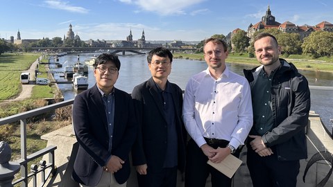 Group photo: four people smiling at the camera. In the background: the Elbe, Carola Bridge and the skyline of the old town with the Frauenkirche, Hofkirche and castle.