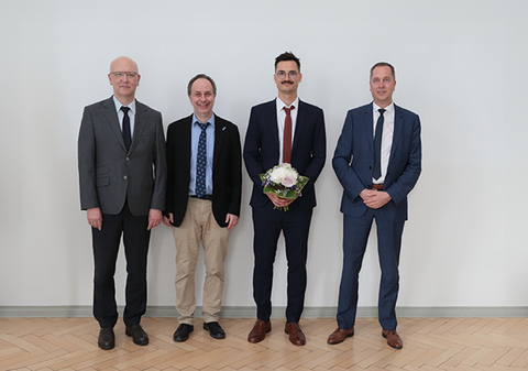 Vier Personen stehen in einer Reihe und schauen in die Kamera. Der Promovend hält einen Blumenstraß in der Hand.