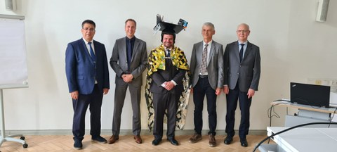 The men are standing next to each other and smiling at the camera. The man in the centre is wearing a doctor's hat.